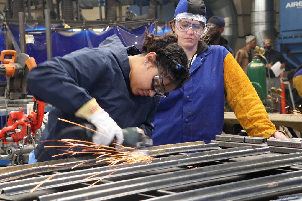 One person grinds metal while another person watches her.